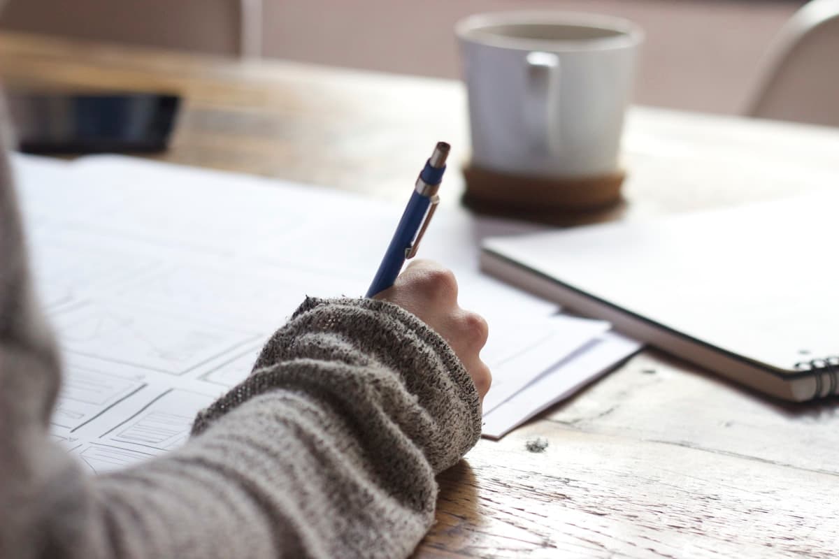 Person writing on paper at a wooden desk with a coffee mug nearby