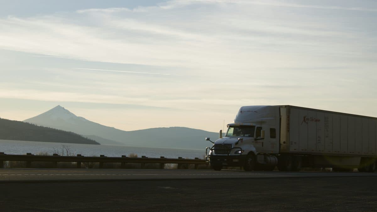 Semi-truck driving along a scenic mountain highway with a lake and volcanic peak in the background