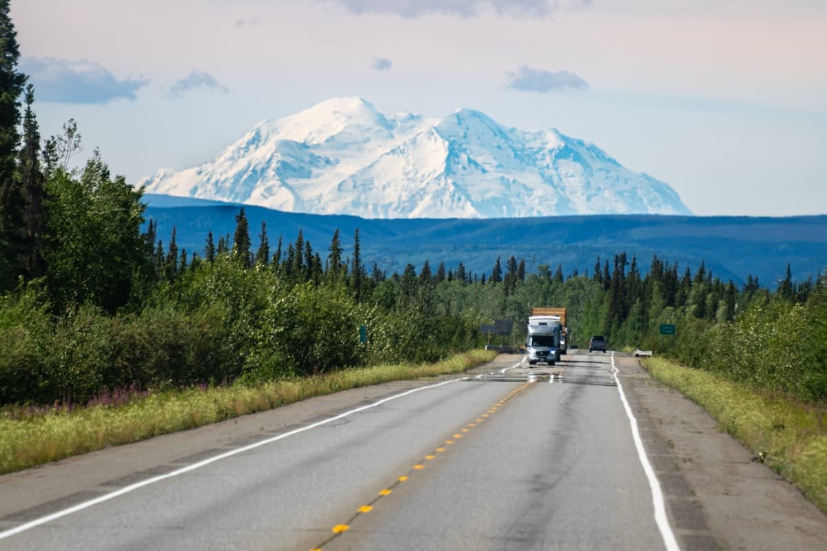 Truck driving down a wide highway with a snow-capped mountain and evergreen forest in the background
